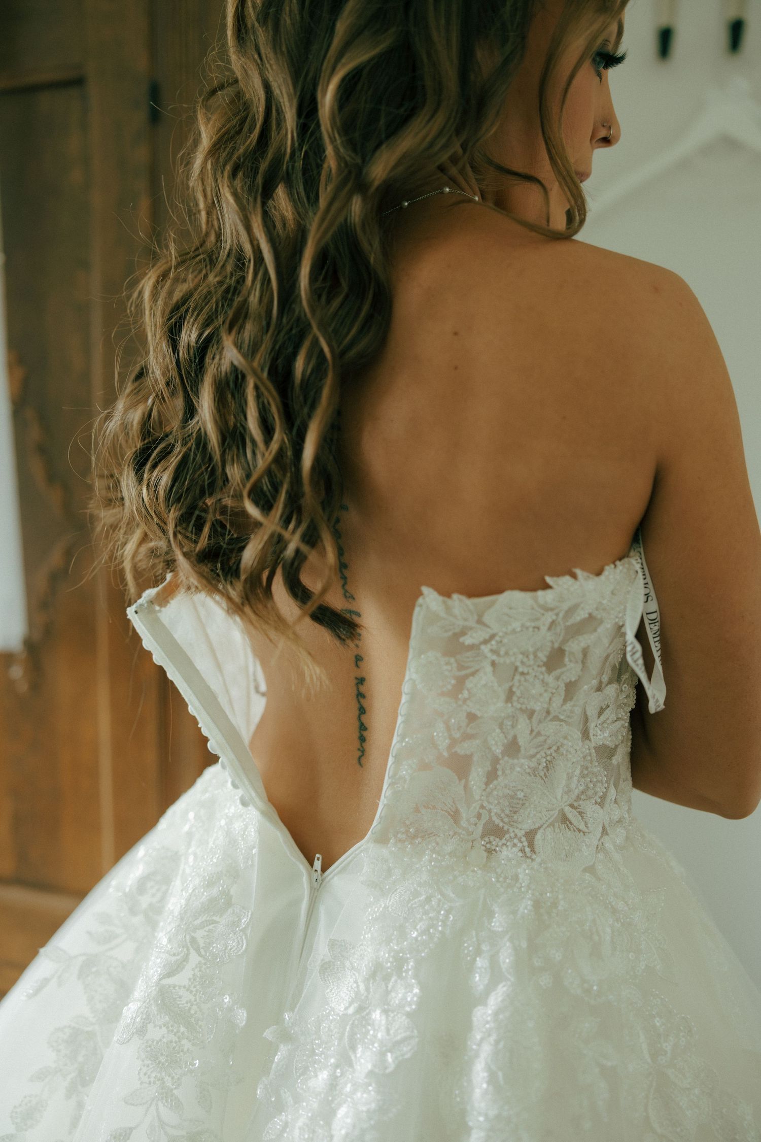 Bride in a strapless lace wedding gown with floral details, showing elegant back design and curly brunette hair.