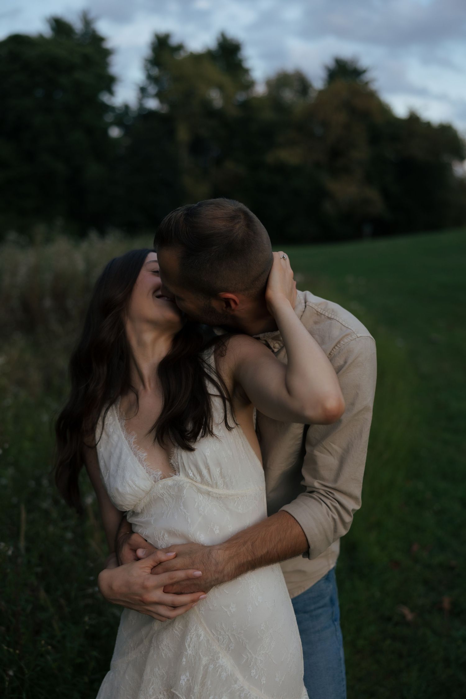 Couple sharing a romantic kiss outdoors at dusk, man in beige shirt embracing woman in white lace dress.