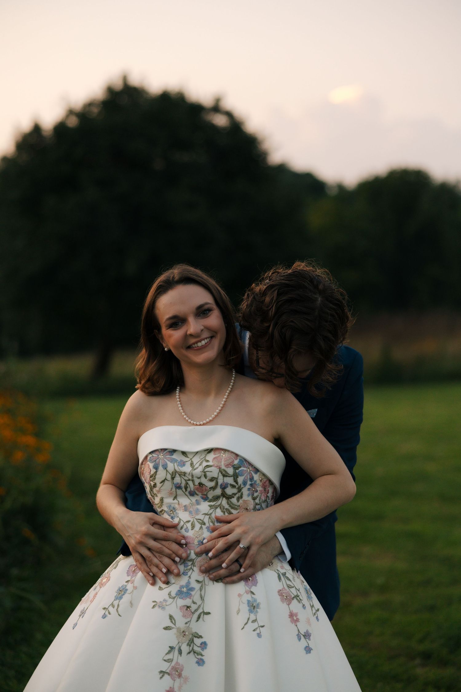 Groom in navy suit embraces smiling bride in strapless floral gown at sunset outdoors.