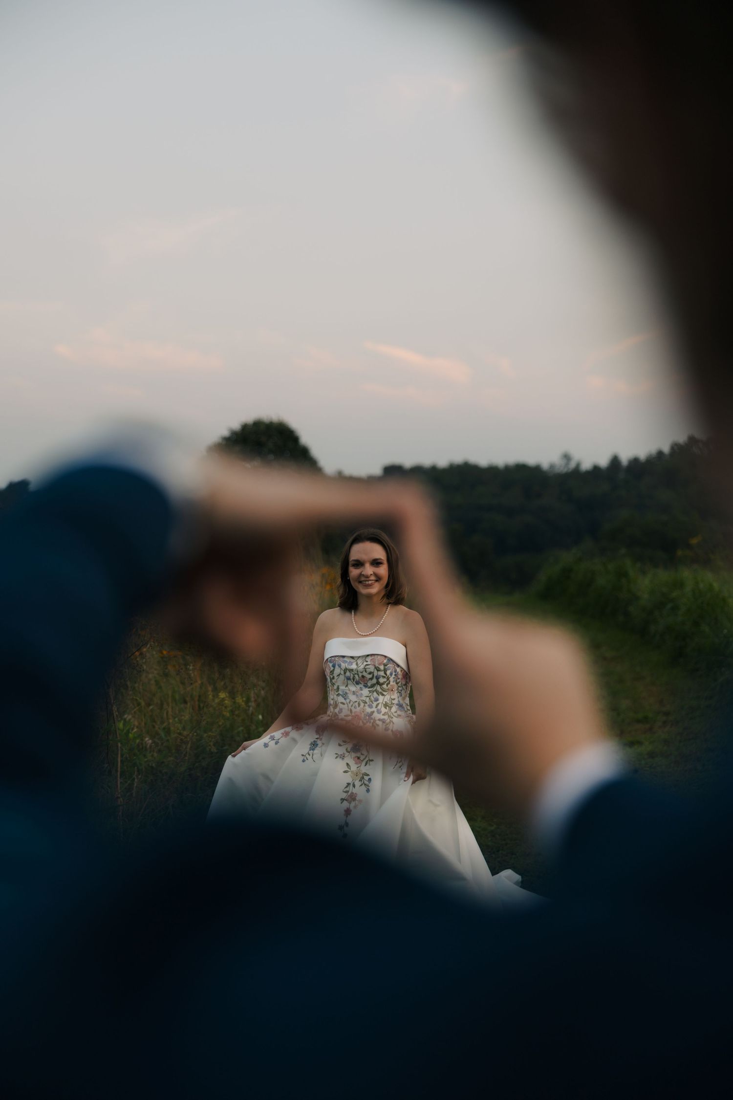 Groom frames bride in his fingers as she smiles in an embroidered wedding gown in a scenic outdoor field.