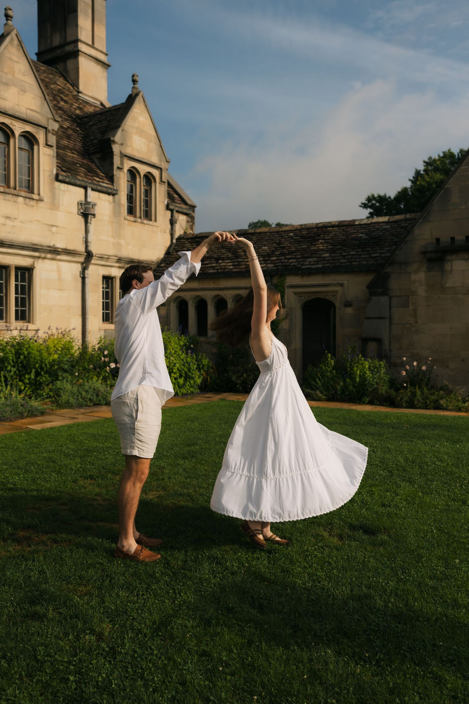 Couple in white outfits dancing on green lawn outside a stone manor house during golden hour.