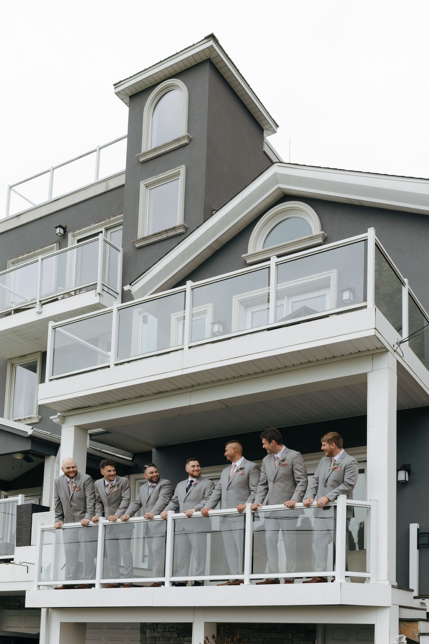 Groomsmen in gray suits stand together on a white balcony of a modern gray building during a wedding.