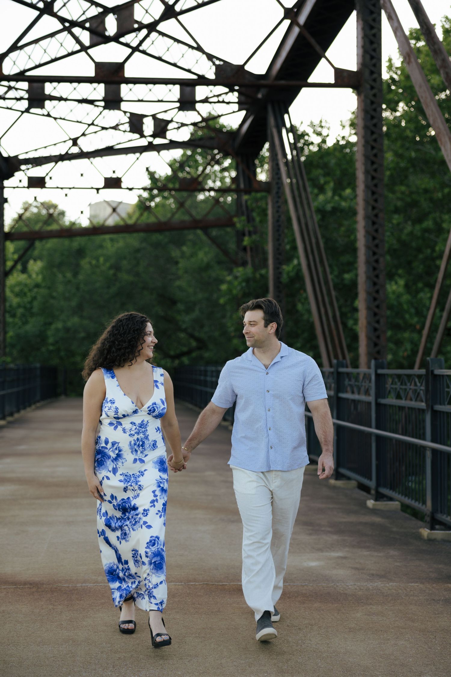 Happy couple holding hands and walking together on a rustic iron bridge surrounded by trees.
