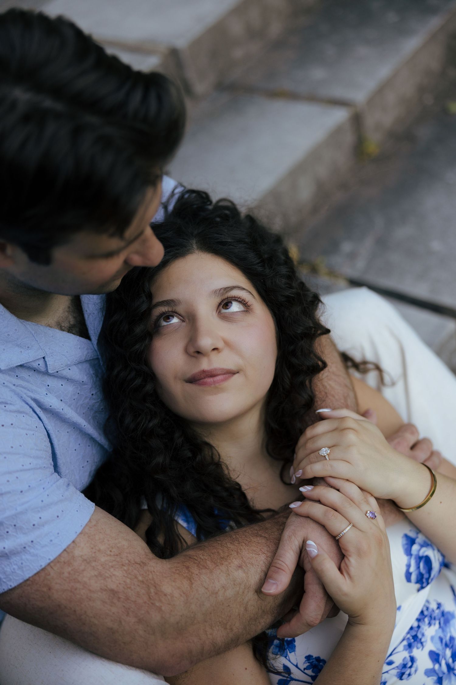 A woman in a blue floral dress looks up lovingly at her partner while resting in his arms on steps.