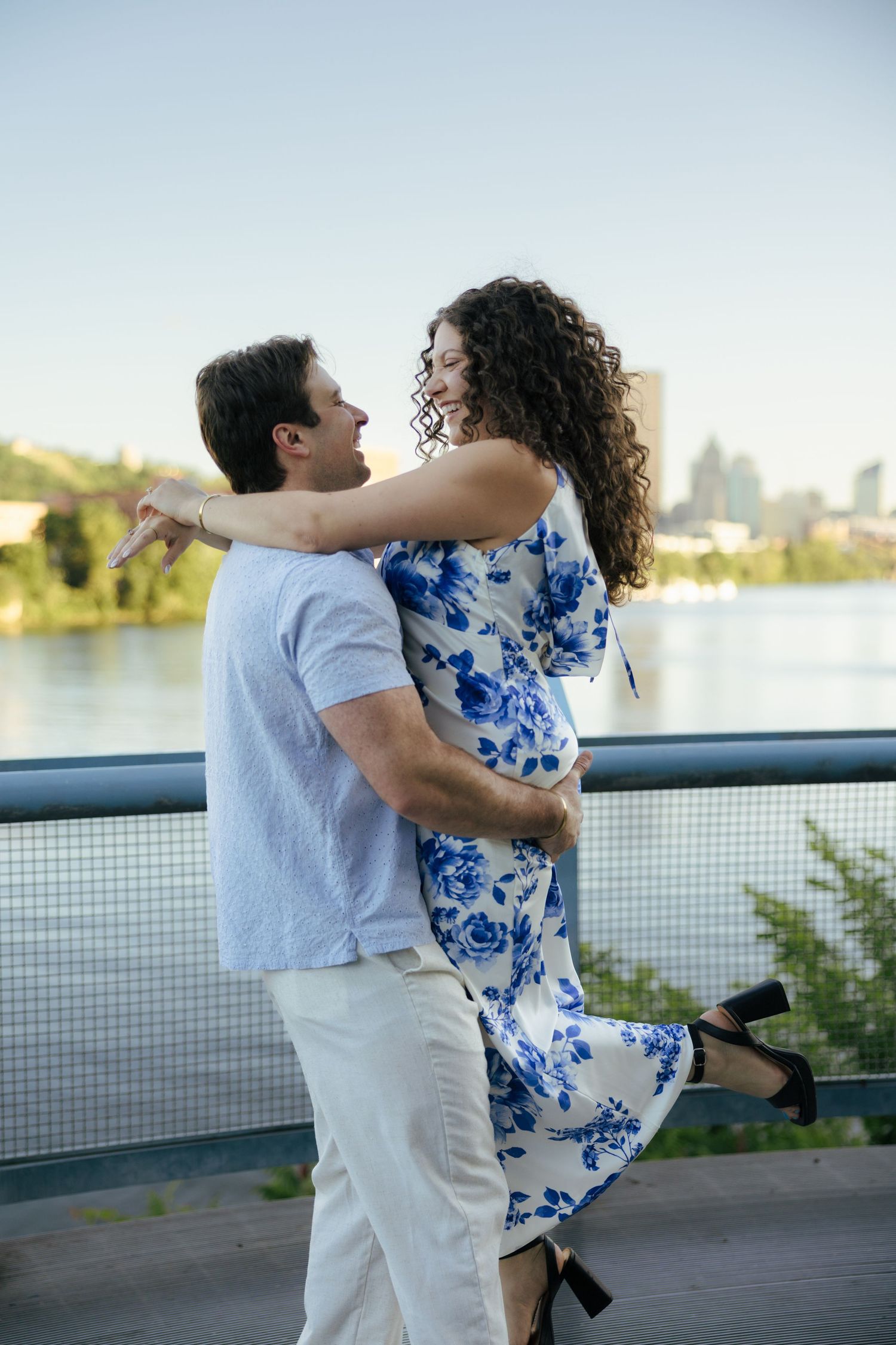 Couple laughing and dancing together on bridge, woman in blue floral dress, river behind them.