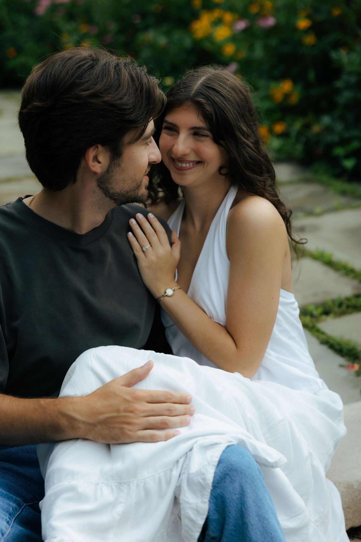 Smiling couple sitting close together outdoors, woman in white dress touching mans chest affectionately.