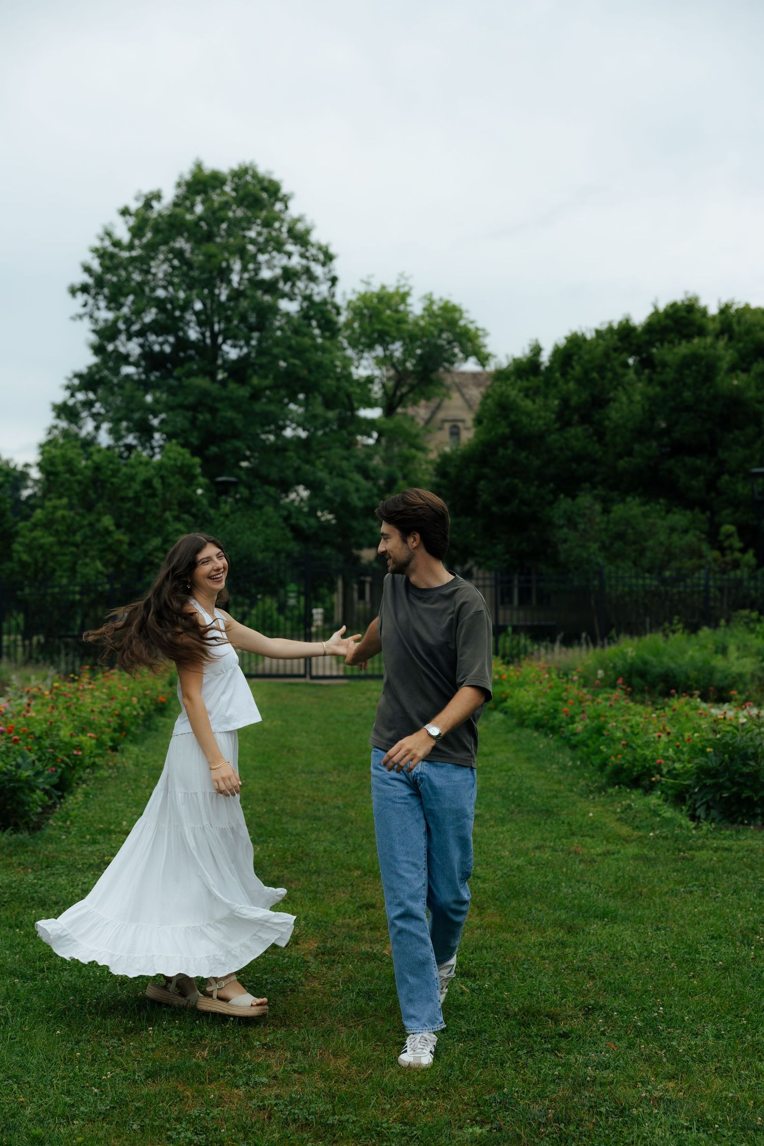 A joyful couple dancing on green grass in a lush garden, woman in white dress, man in gray tee and jeans.