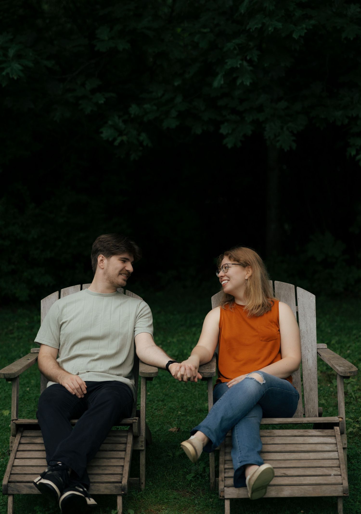 Smiling couple holding hands while seated in wooden Adirondack chairs in a shaded garden.