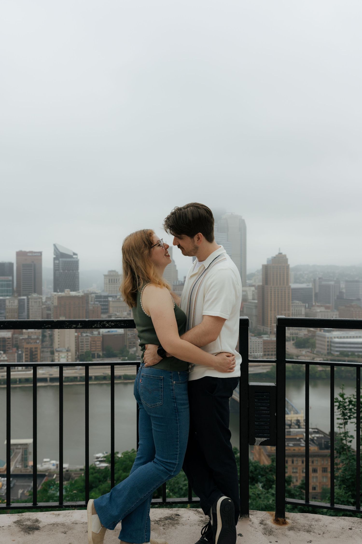 Romantic couple holding each other close on a scenic overlook with Pittsburgh skyline visible behind them.