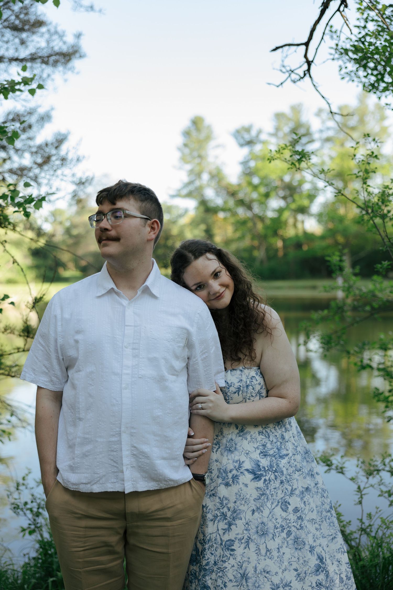 Couple poses by a serene pond surrounded by lush green trees, woman in floral dress leans on mans shoulder.