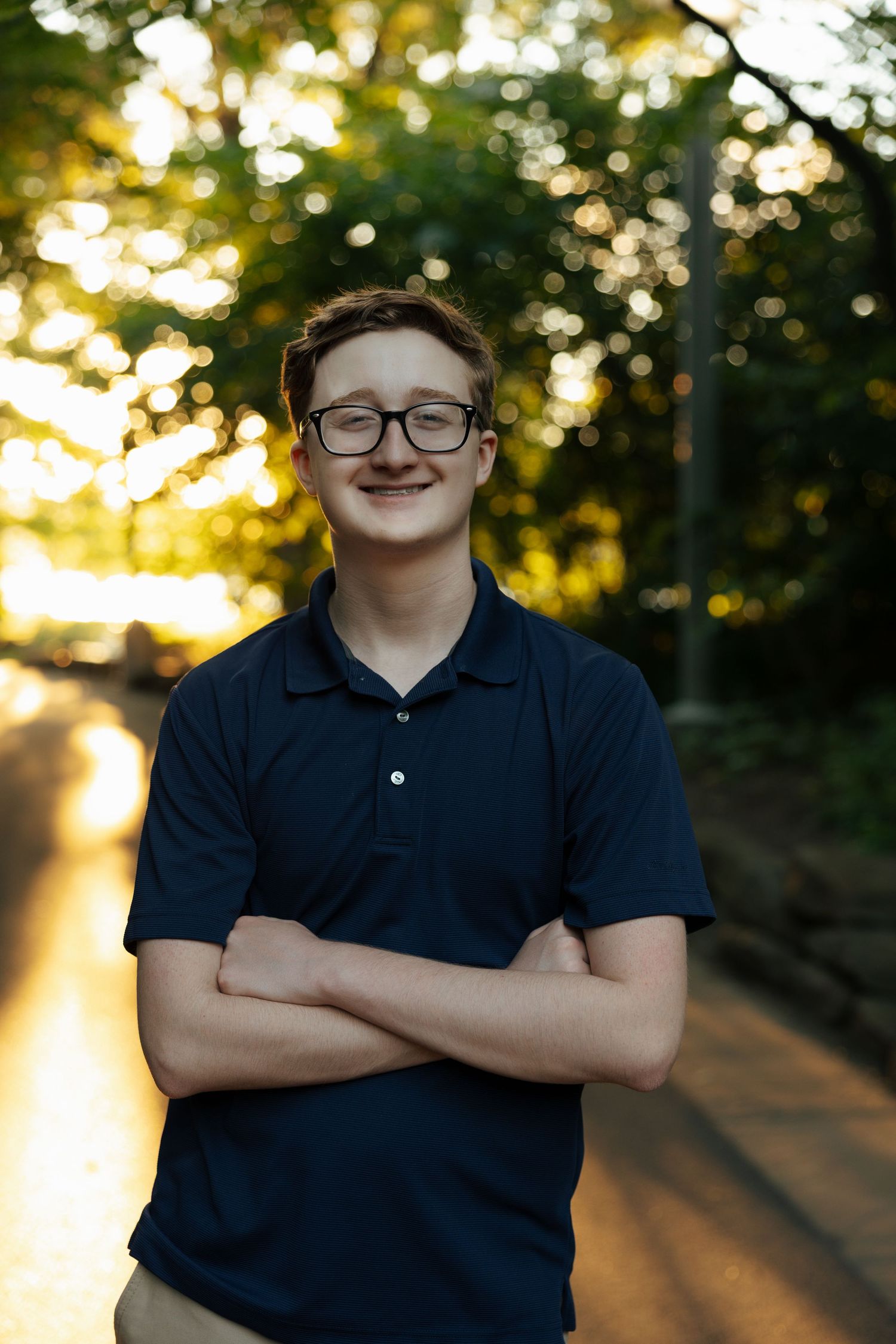 Smiling teenage boy in navy polo with arms crossed standing on sunlit path surrounded by green trees.