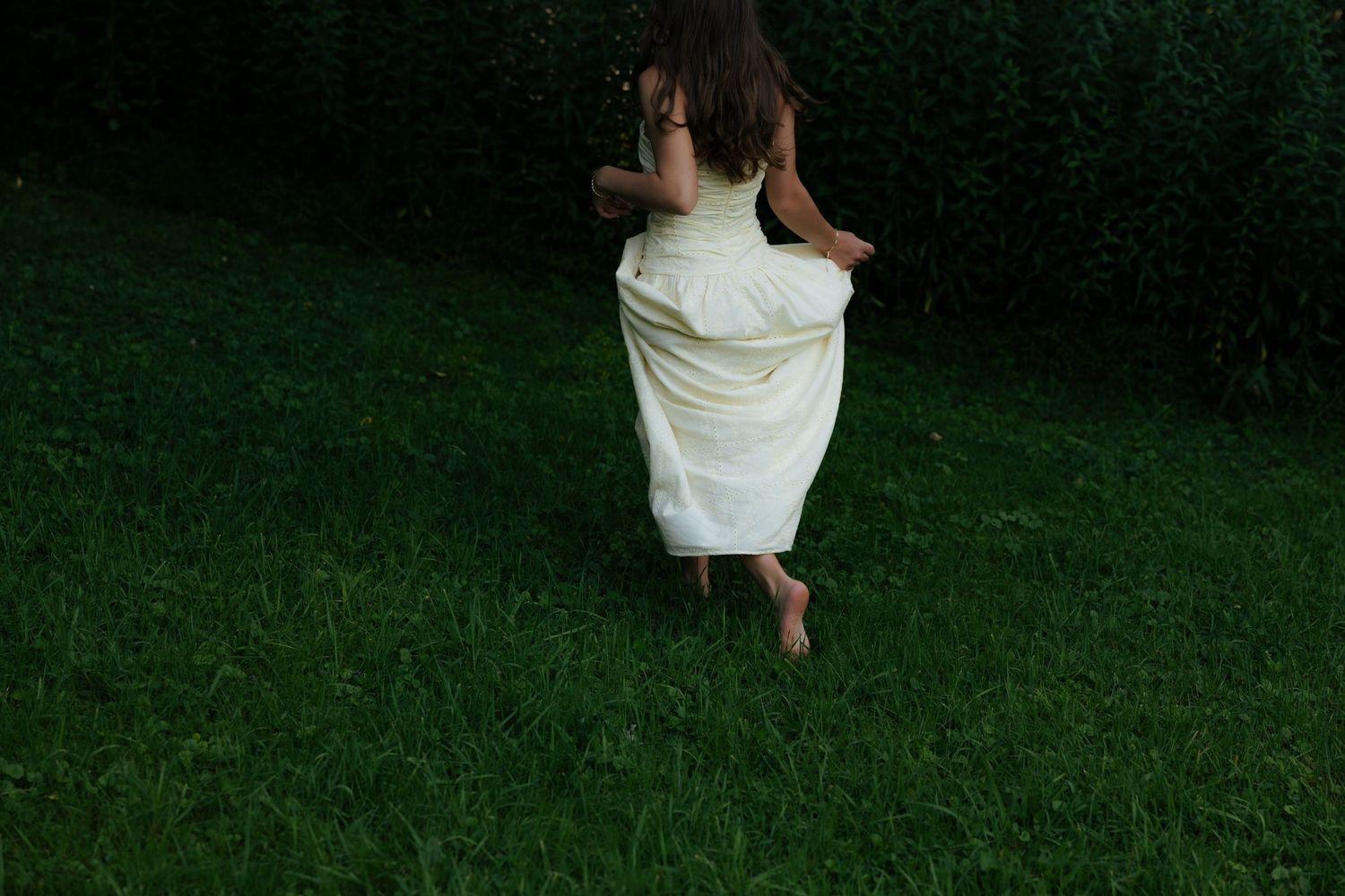 A barefoot girl in a white dress runs across lush green grass, captured from behind in motion.