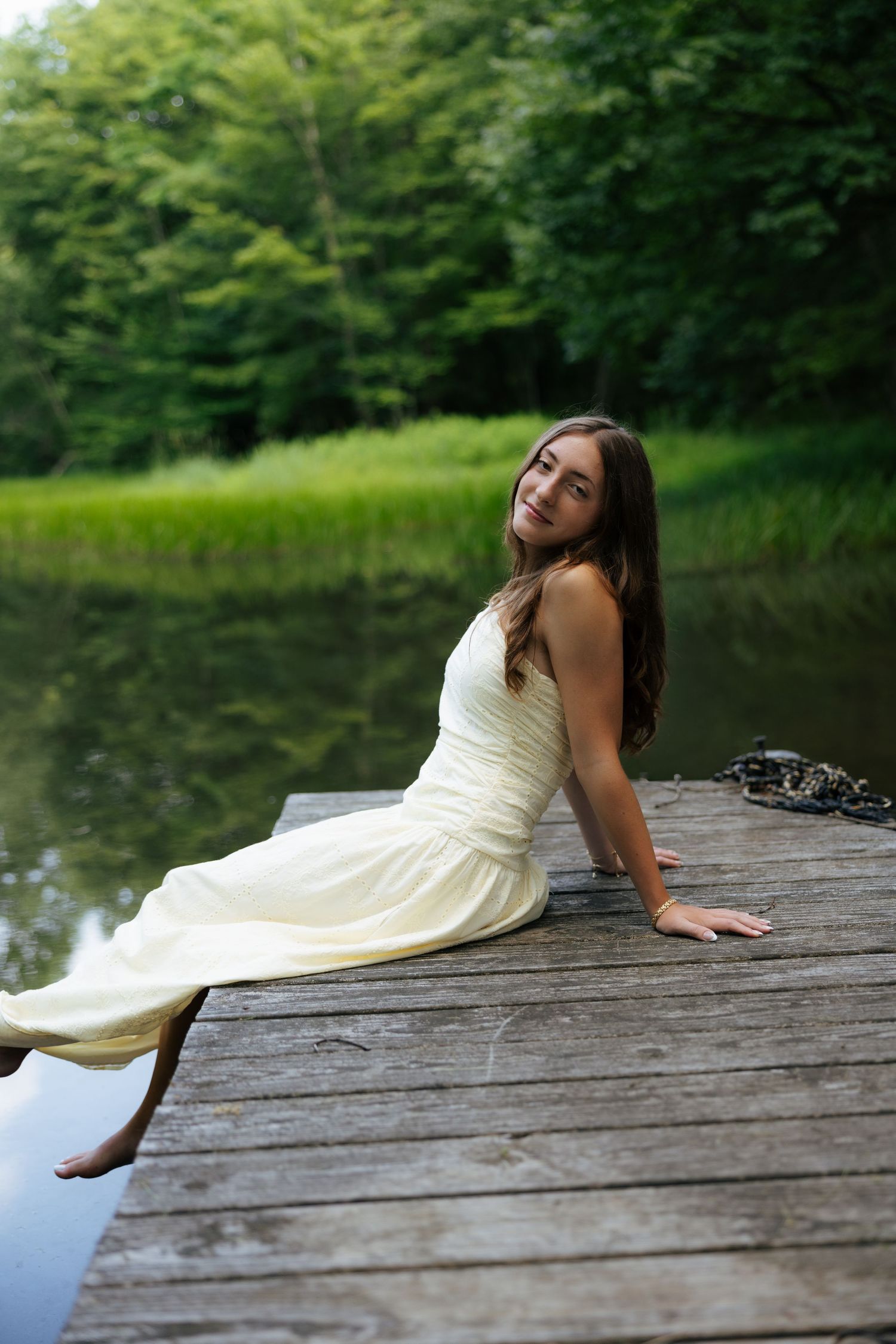 Young woman in cream dress sitting on wooden dock by calm lake surrounded by trees.