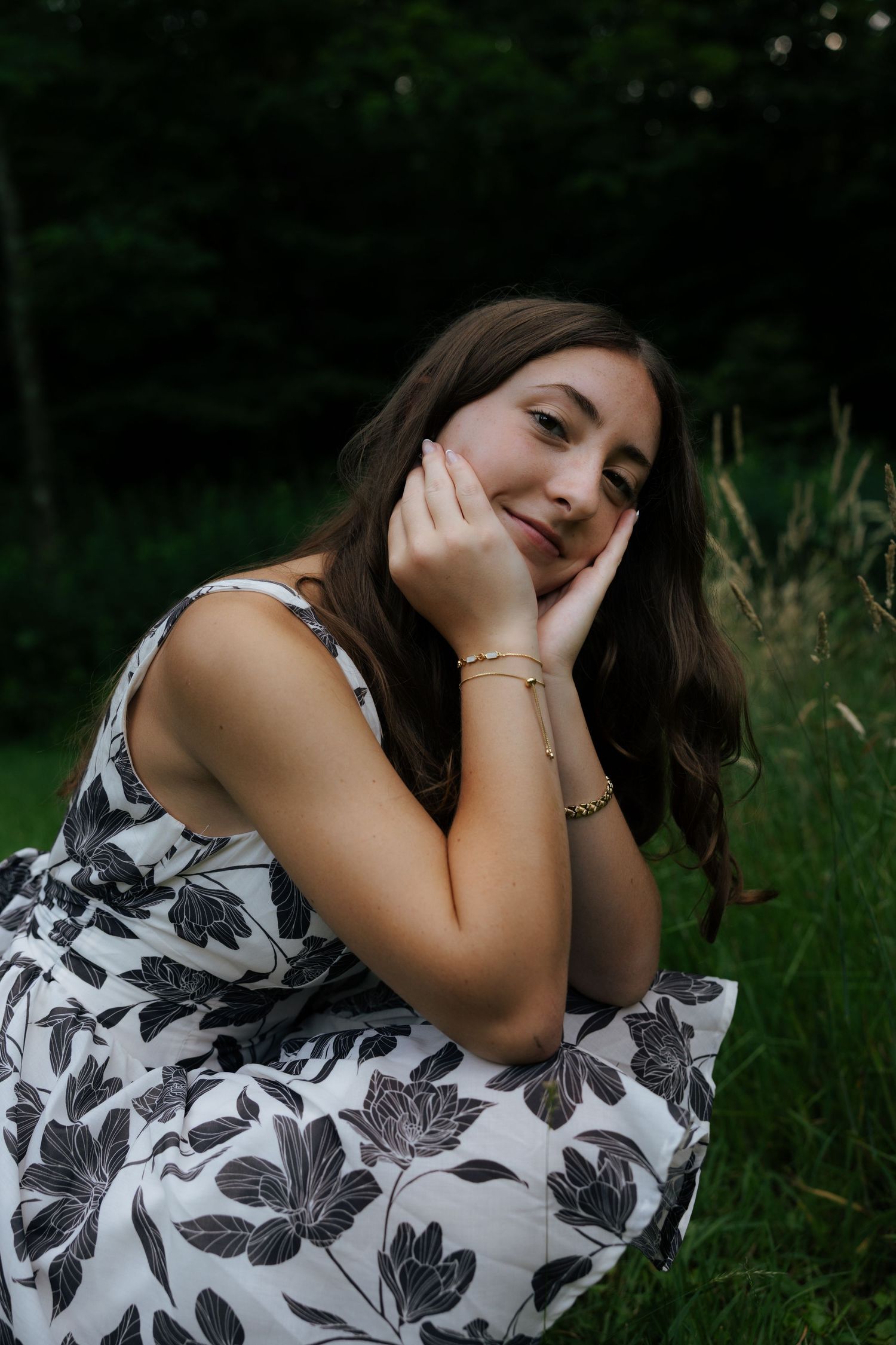 Brunette woman in floral dress sitting in grass with hands on cheeks.