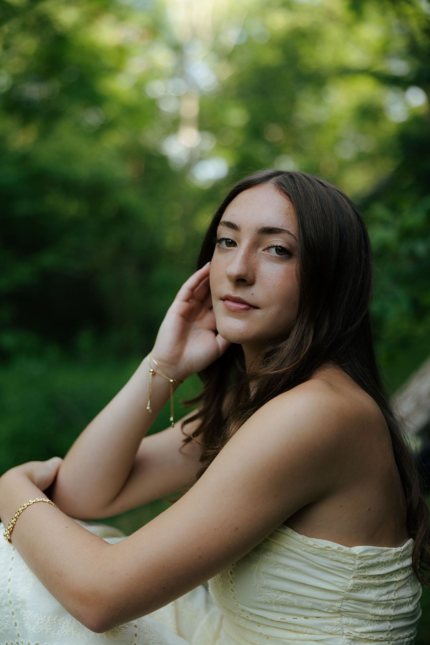 Beautiful young woman in white strapless dress sitting outdoors in lush green forest setting.