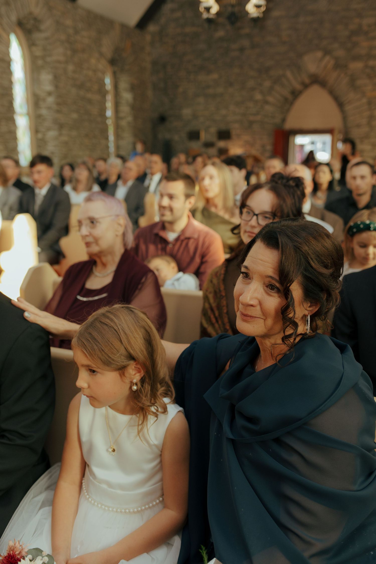 Wedding guests seated in historic stone church watching ceremony, woman in teal wrap with young flower girl in front.