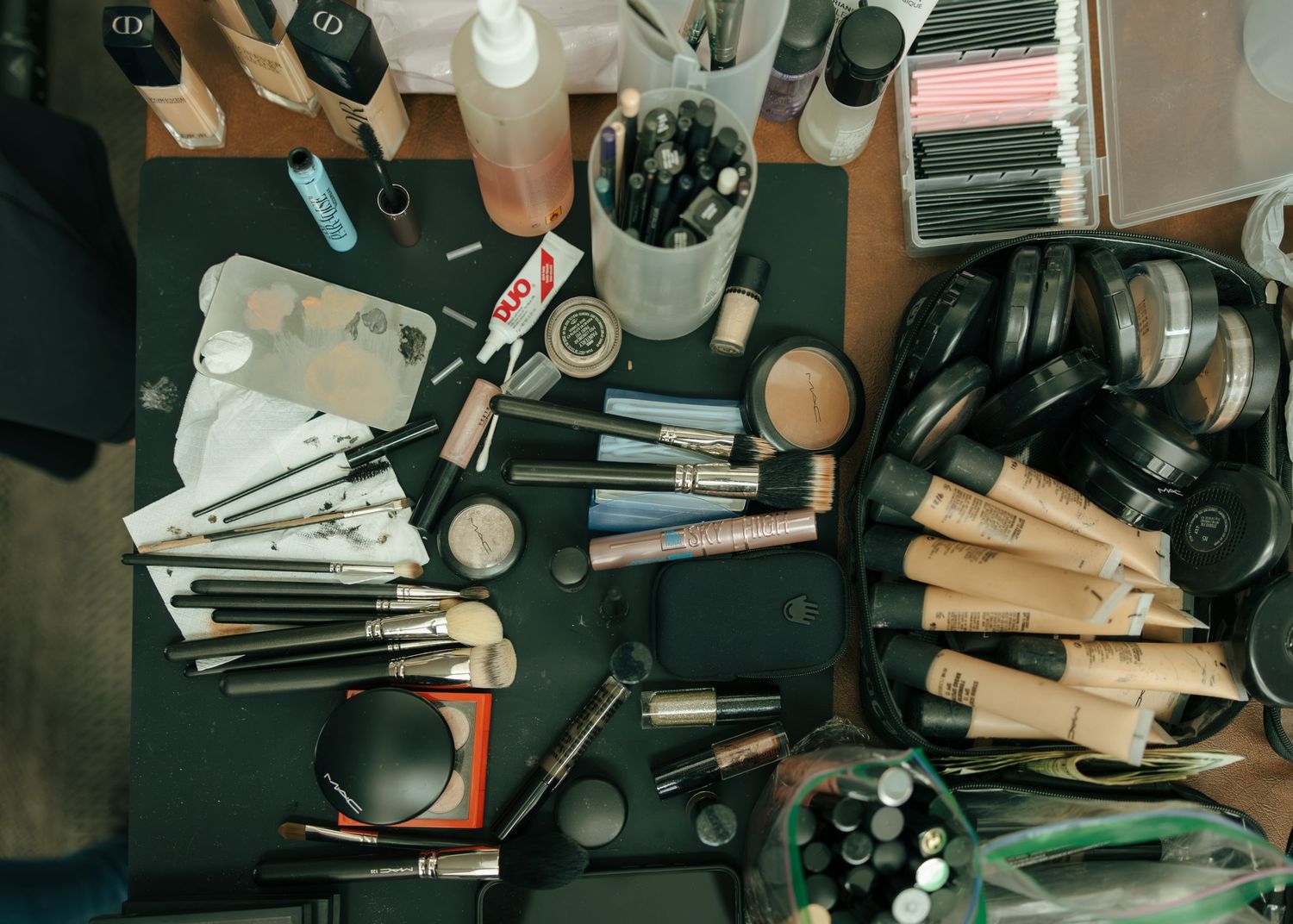 Cluttered makeup vanity with brushes, foundations, eyeshadows, and beauty tools spread across a dark surface.