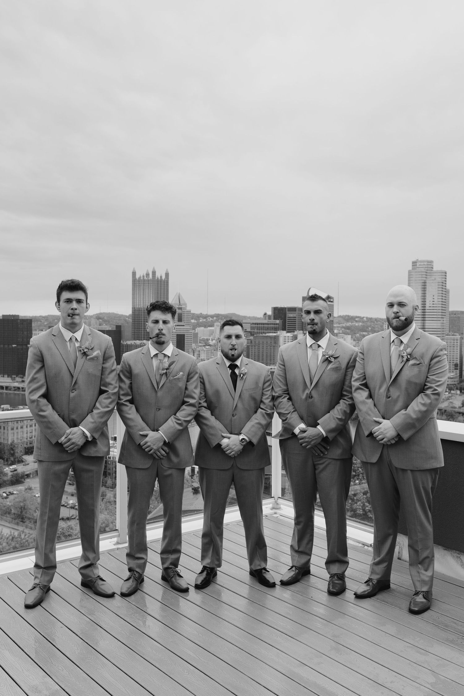 Black and white photo of five groomsmen in suits posing on a rooftop with a city skyline backdrop.