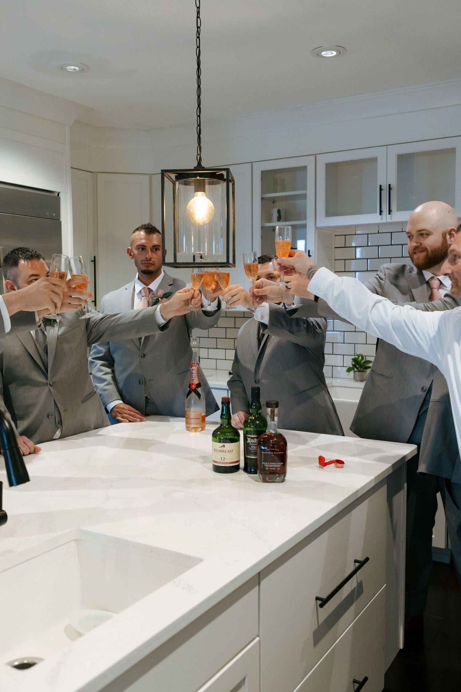 Groomsmen in gray suits toasting with amber drinks around a white marble kitchen island with whiskey bottles.