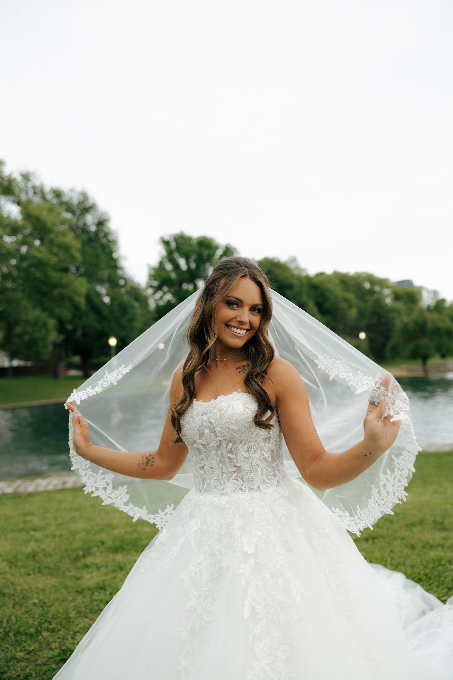 Smiling bride in lace strapless wedding gown holding veil outdoors near a pond with green trees in background.