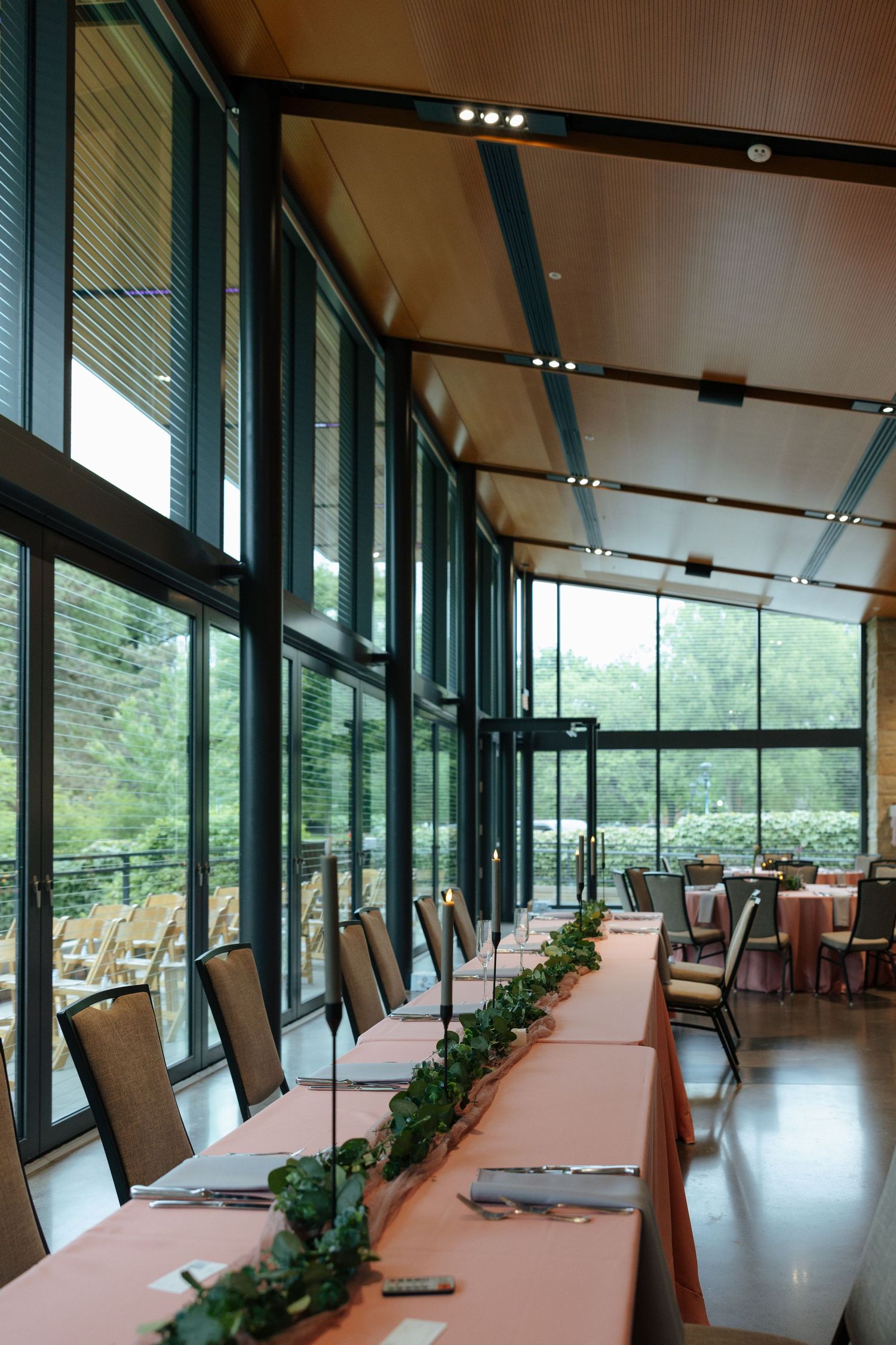Elegant banquet hall with floor-to-ceiling windows, long tables with pink linens and greenery centerpieces overlooking lush gardens.