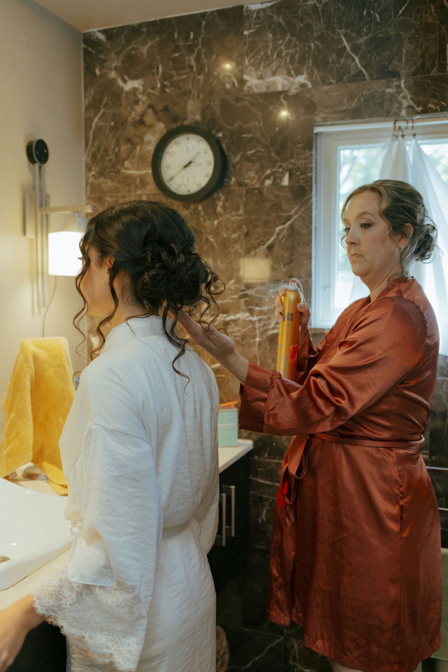 Bridesmaid in orange satin robe sprays hairspray on bride's updo in marble bathroom before wedding.