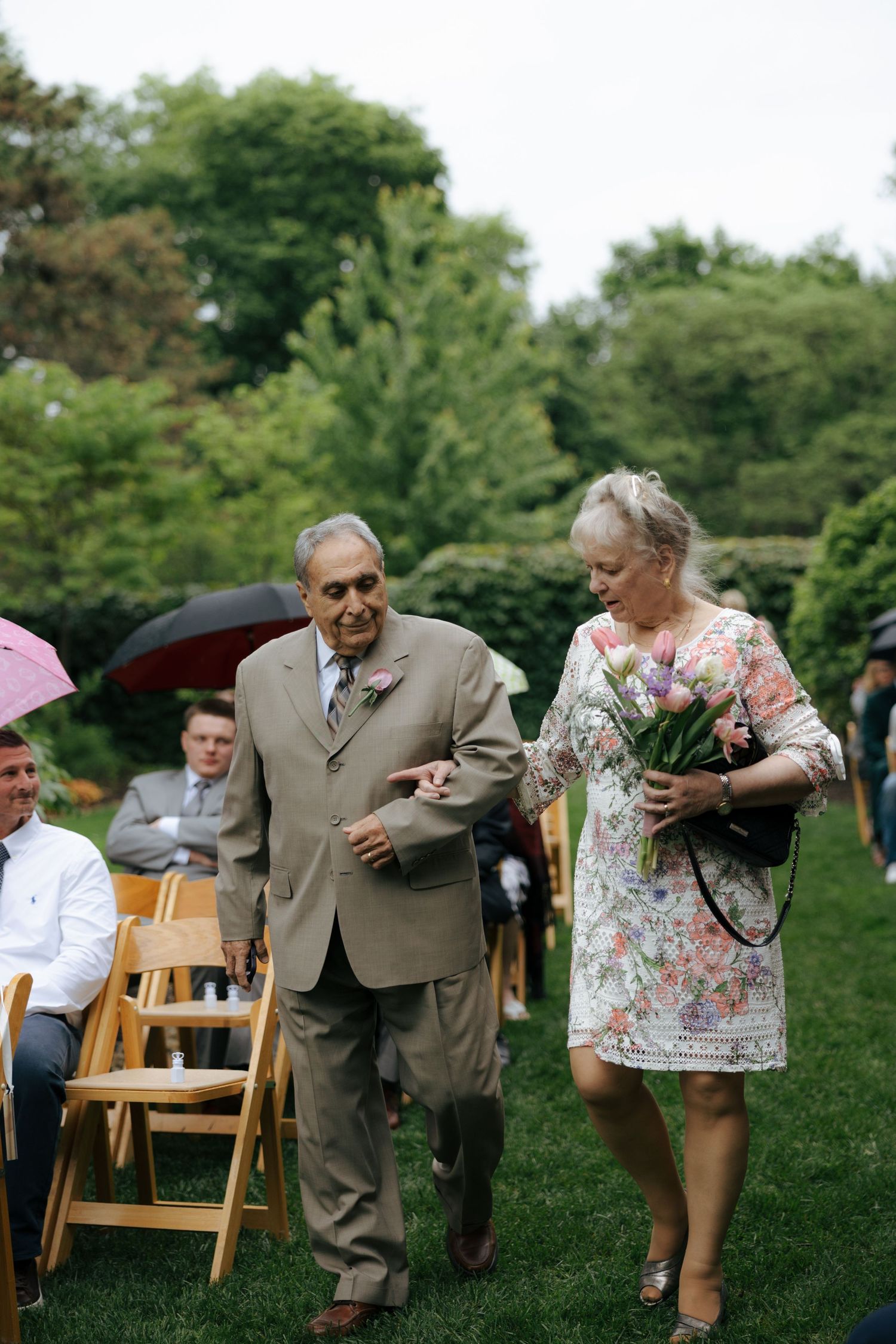 Elderly couple walking down outdoor wedding aisle, woman holding colorful bouquet, guests seated on wooden chairs in garden.