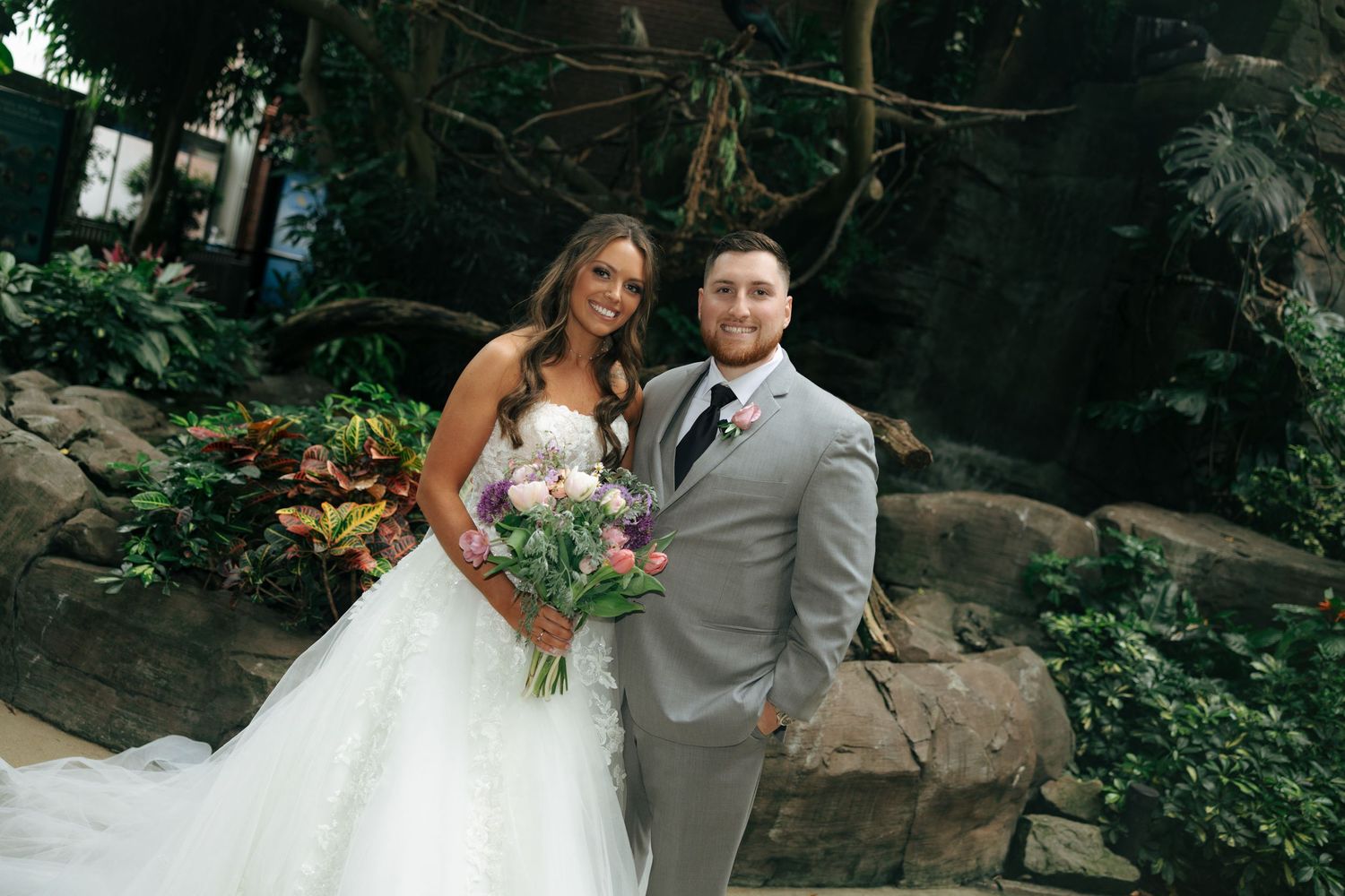 Bride and groom posing together in a lush garden setting, bride holding a colorful bouquet in a white gown.