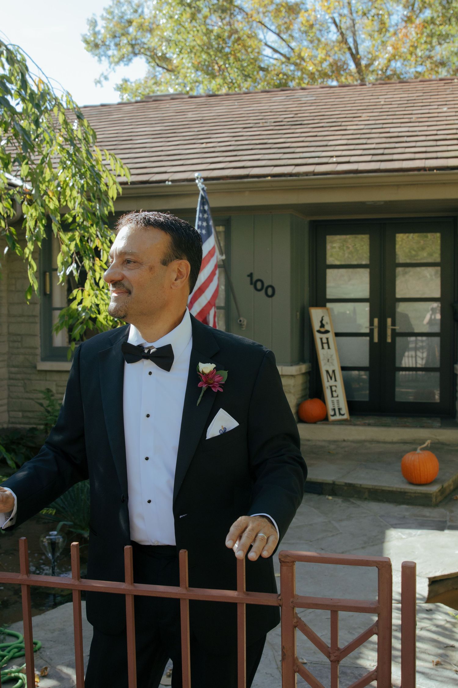 Groom in black tuxedo with boutonniere smiling outside home with American flag on wedding day.