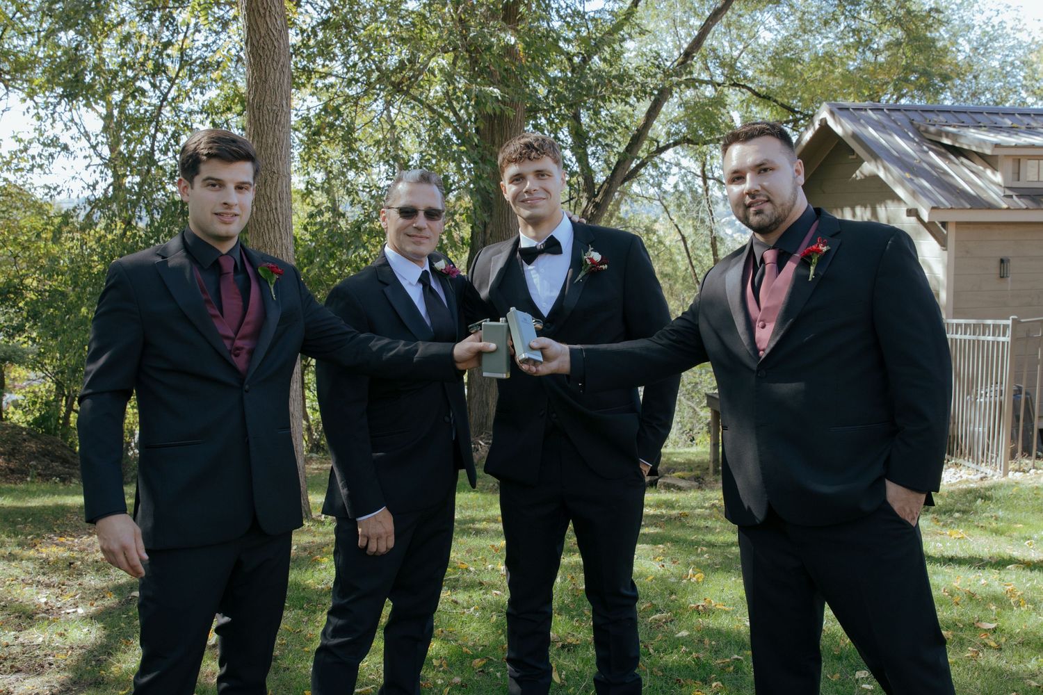 Four young men in black suits with red accessories posing outdoors for a formal prom or wedding photo.