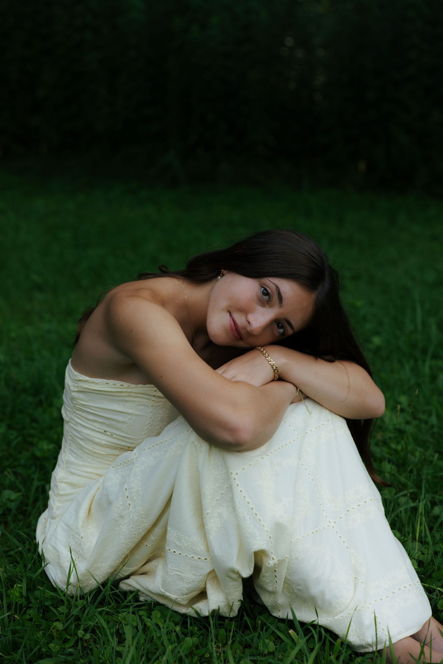 Young woman in white dress sitting on green grass, hugging her knees and smiling softly at camera.