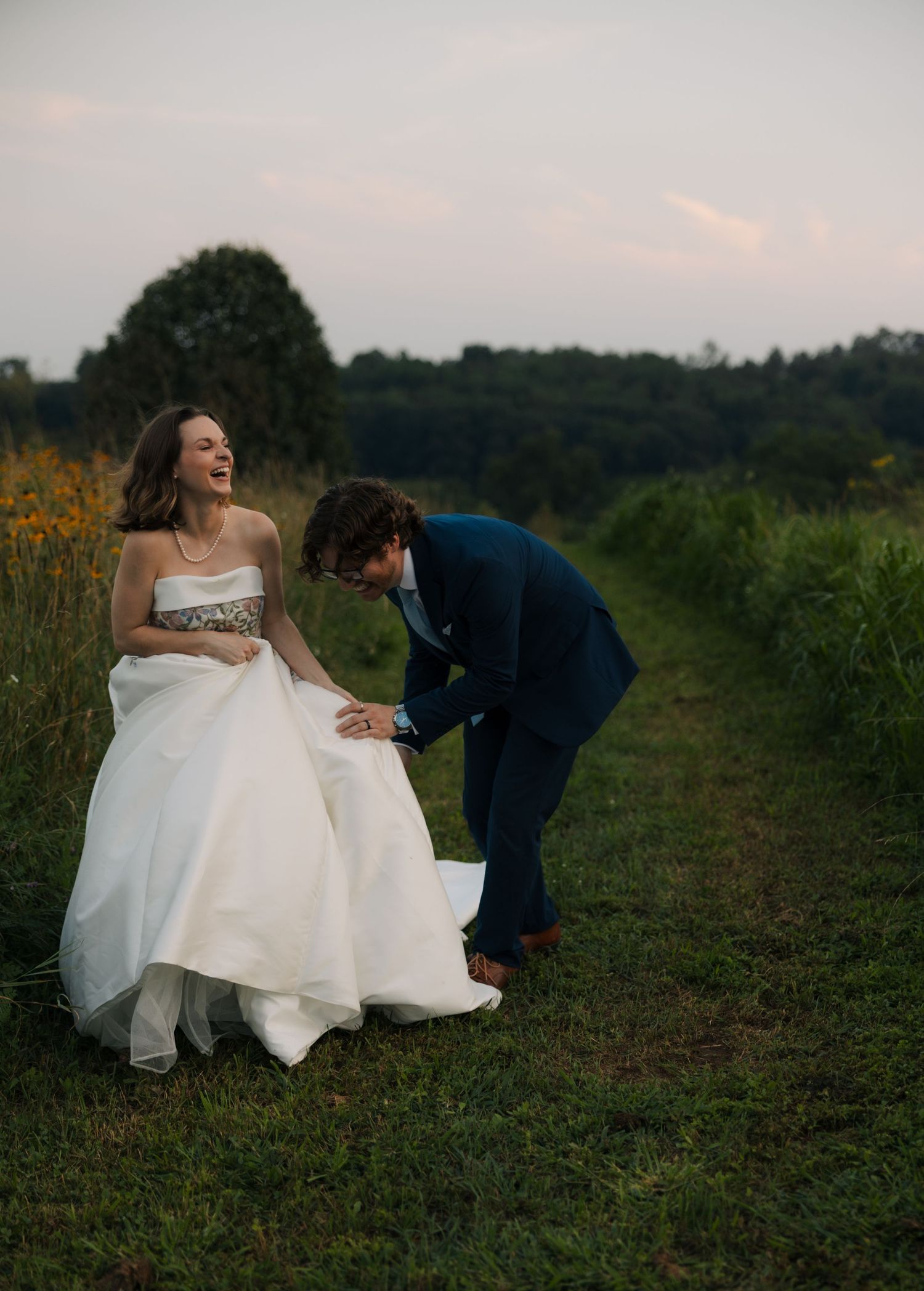 A bride in a white gown laughs joyfully as a groom in a navy suit bows toward her in a lush green field at dusk.