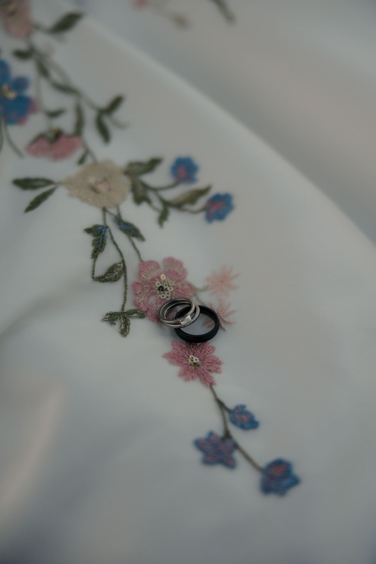 Wedding rings resting on delicate floral embroidery of a white bridal gown.