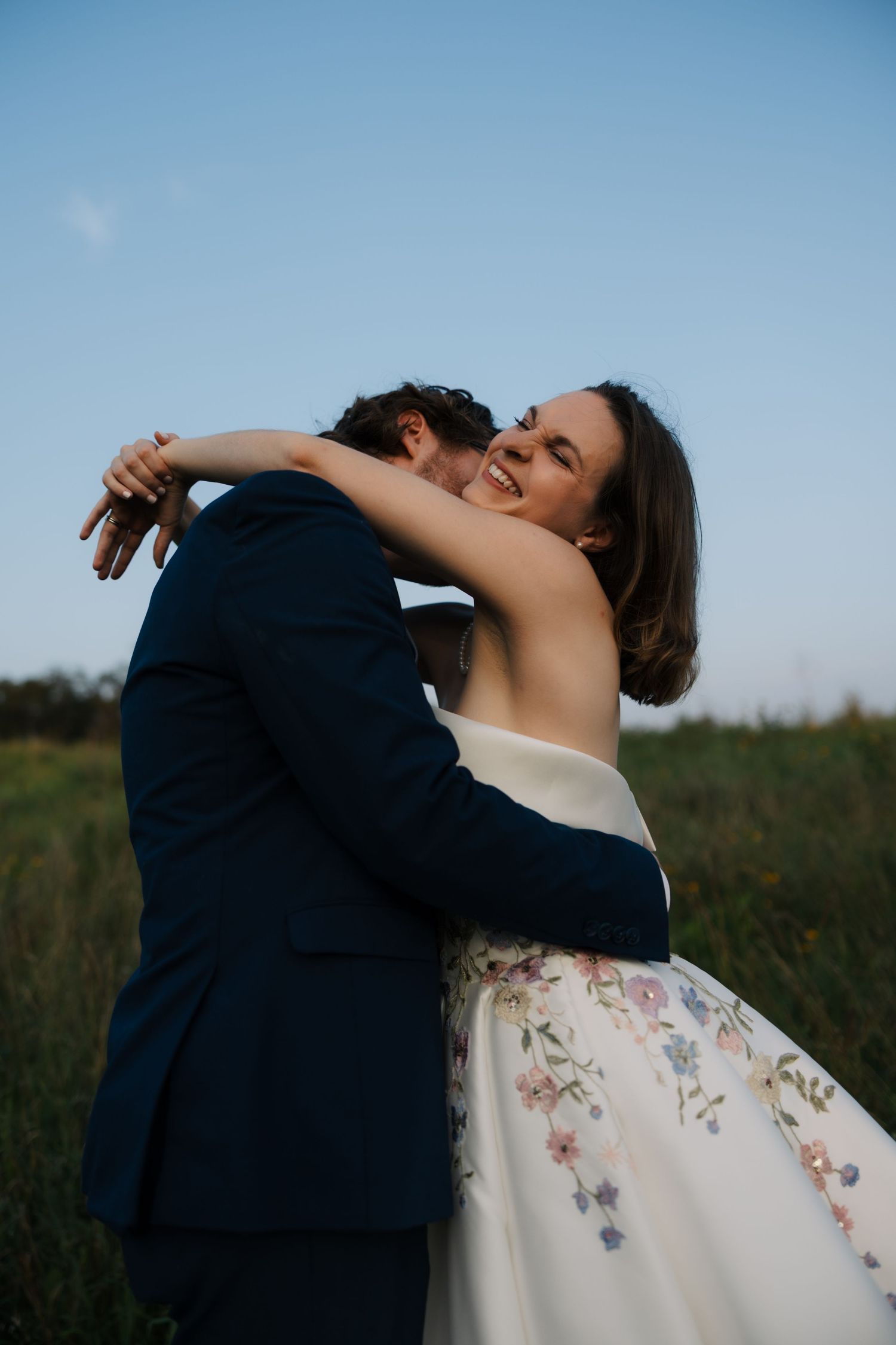Smiling bride in floral strapless gown embraces groom in navy suit in a sunlit outdoor field.
