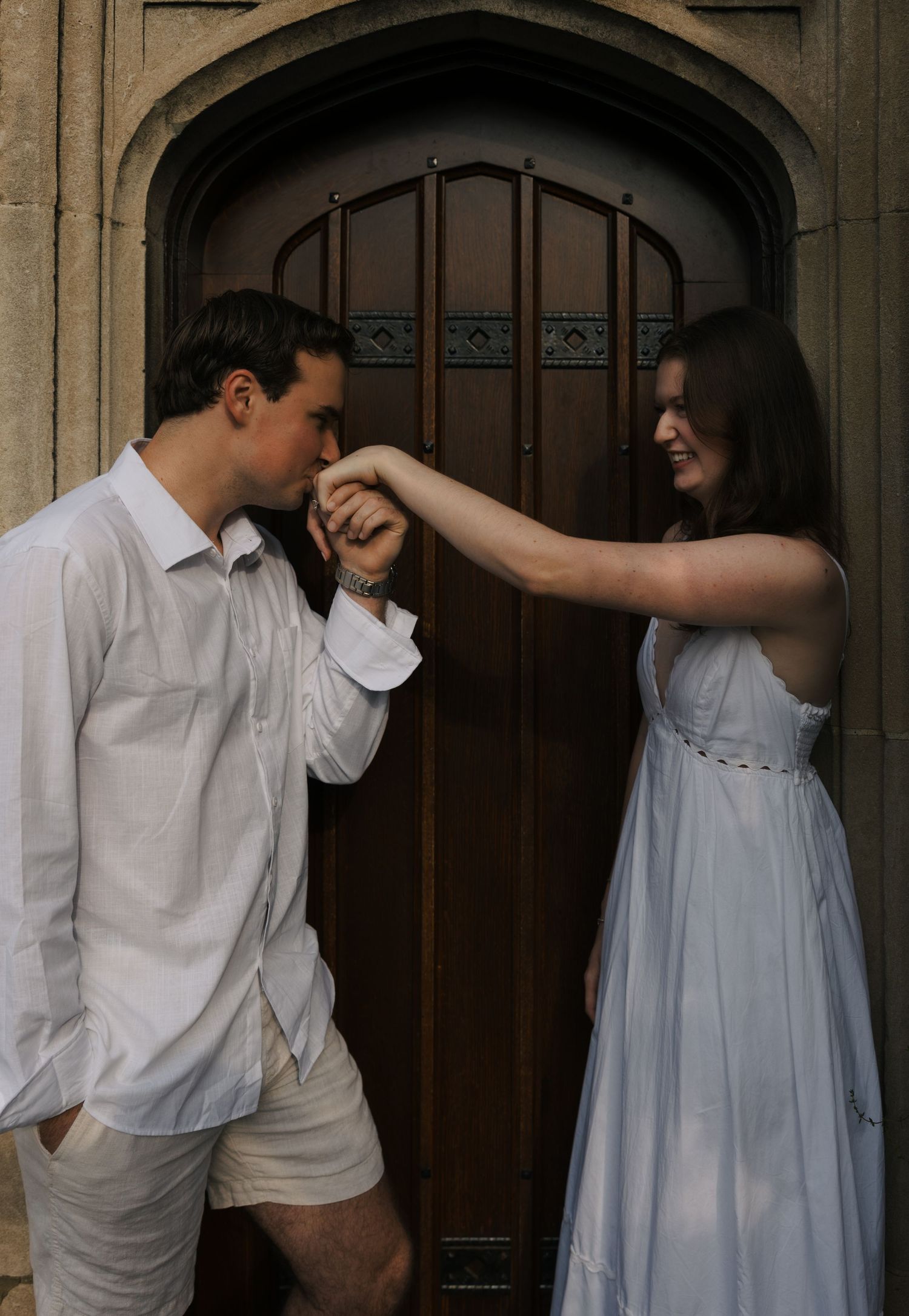 A man in white shirt kisses a woman's hand as she smiles in a light blue dress before an arched wooden door.