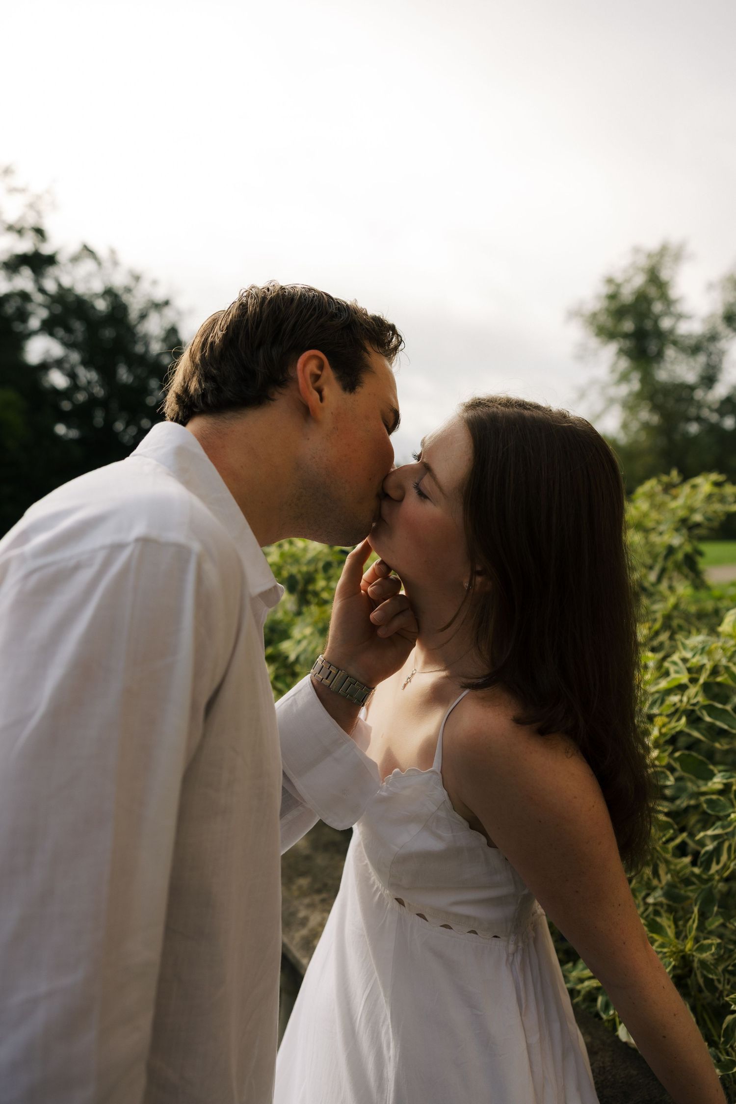 Couple sharing a gentle kiss outdoors surrounded by lush green foliage at dusk.