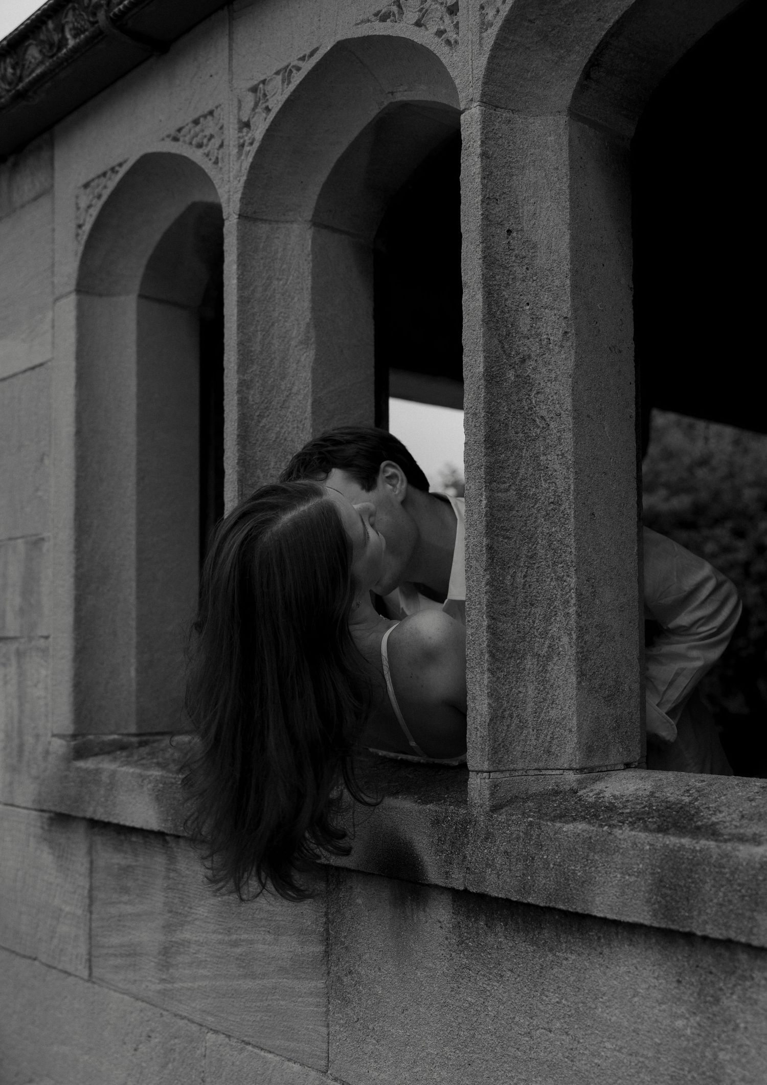 A couple shares a kiss through a stone archway window in a dramatic black and white romantic portrait.