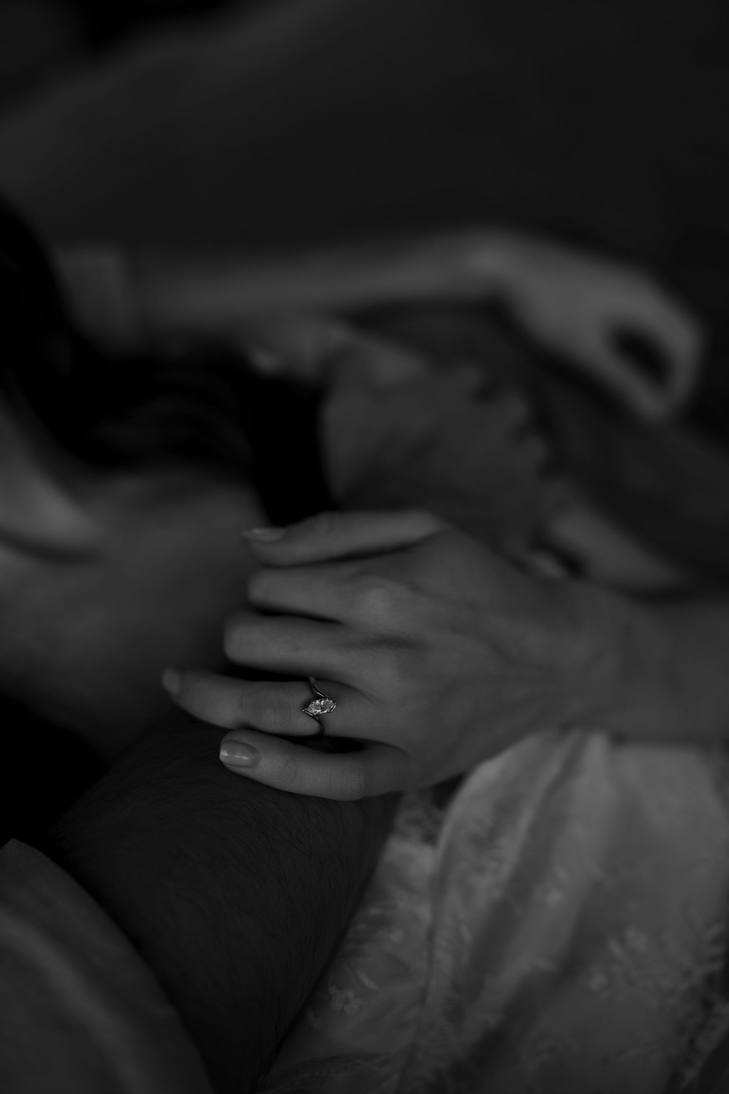 Black and white photo of two hands intertwined, one wearing a diamond engagement ring, resting on a white lace dress.