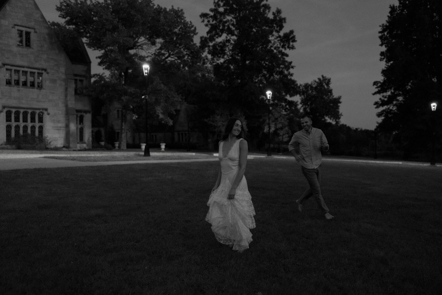 Black and white photo of couple walking on lawn at night near illuminated stone estate building.