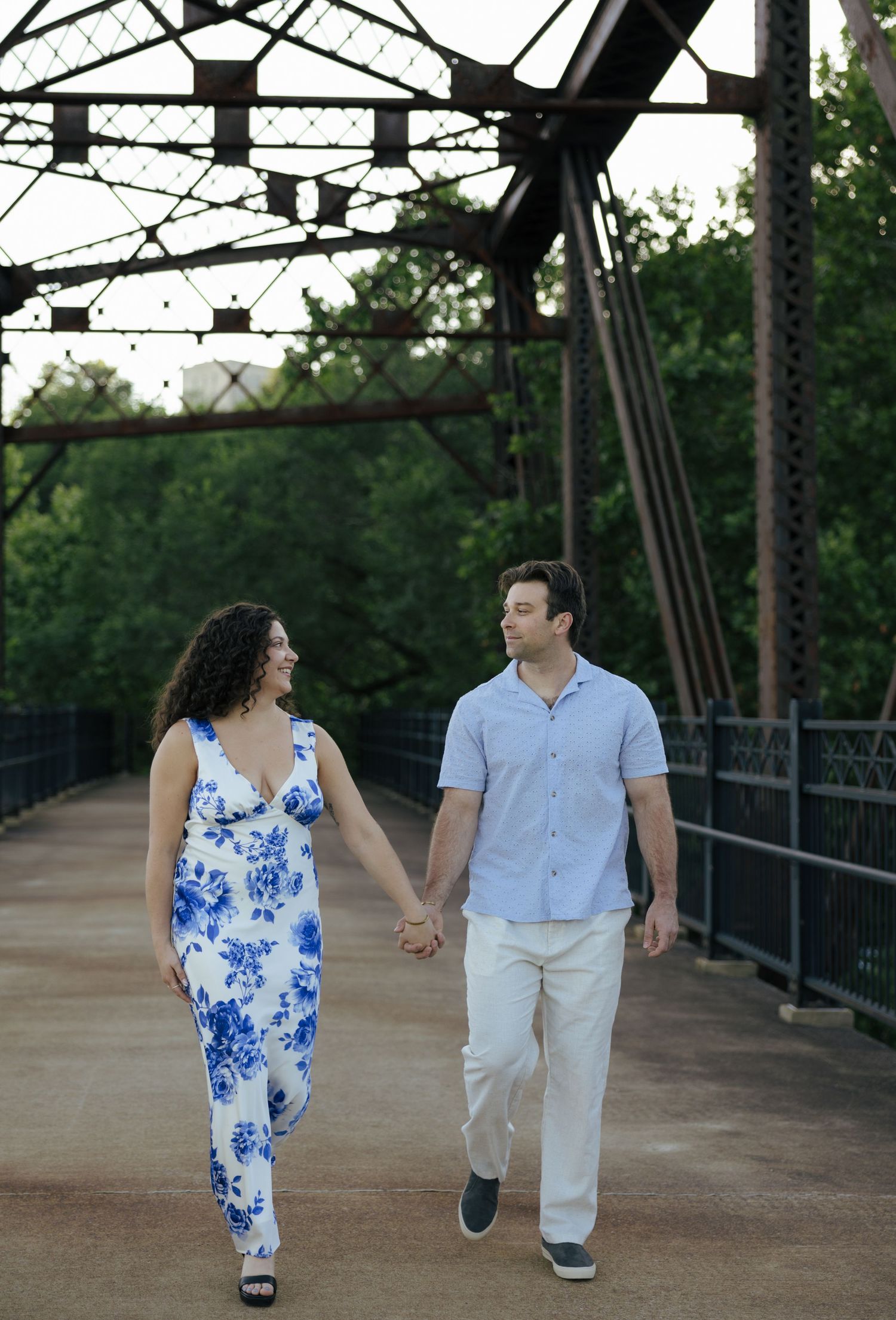 Engaged couple holding hands and walking across a historic iron bridge surrounded by lush green trees at sunset.
