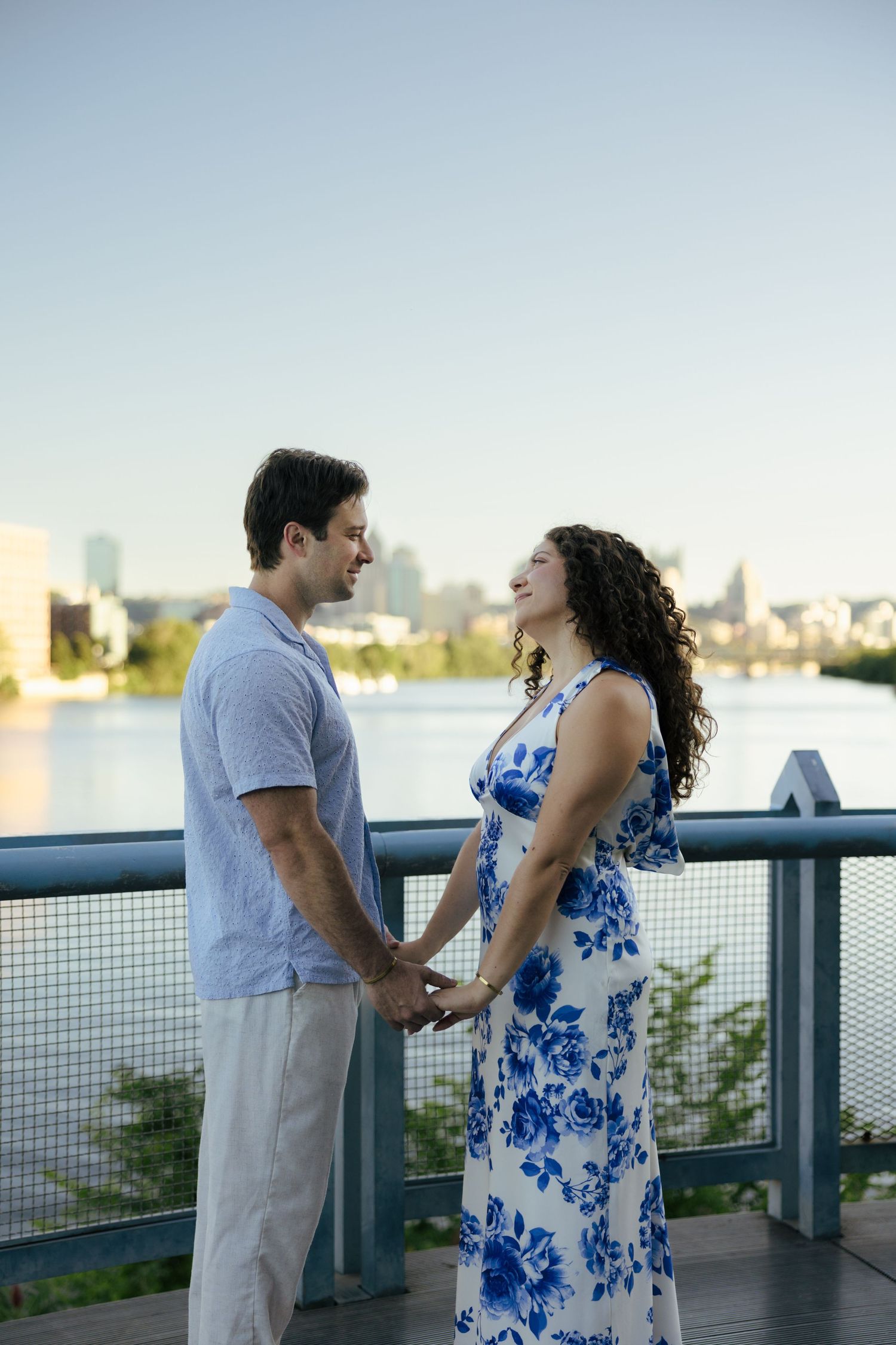 Smiling couple holding hands on bridge overlooking river with urban skyline at golden hour.