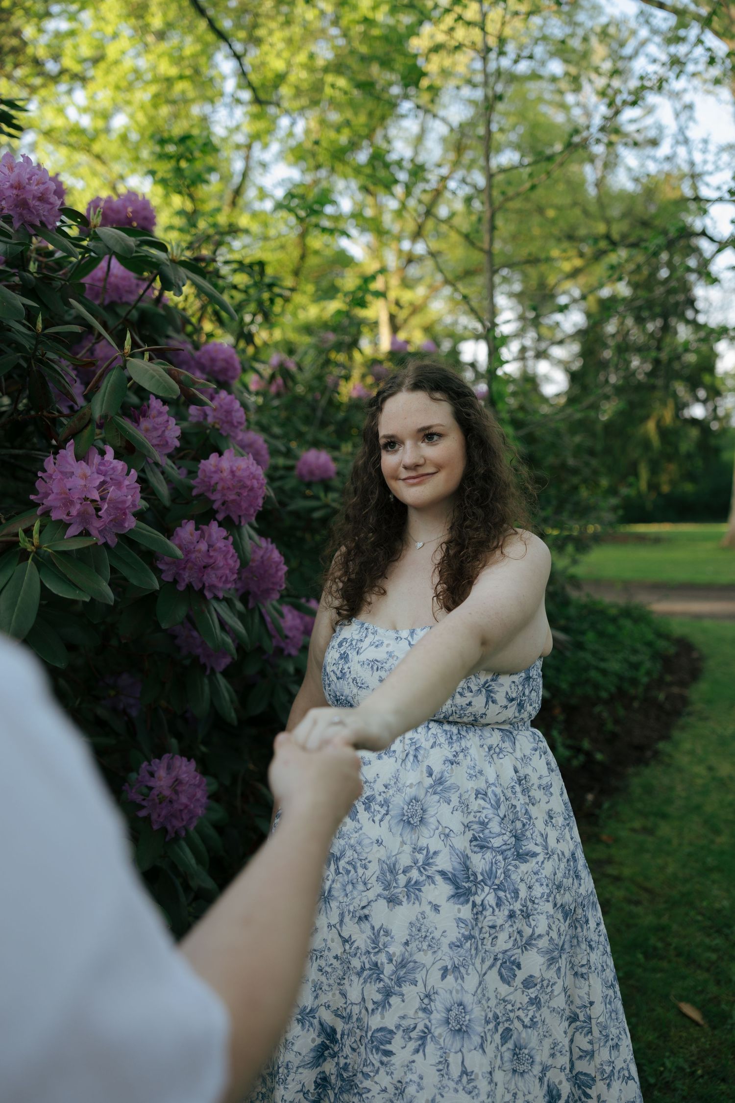 Young woman in blue floral dress holds mans hand near blooming purple rhododendrons in park.