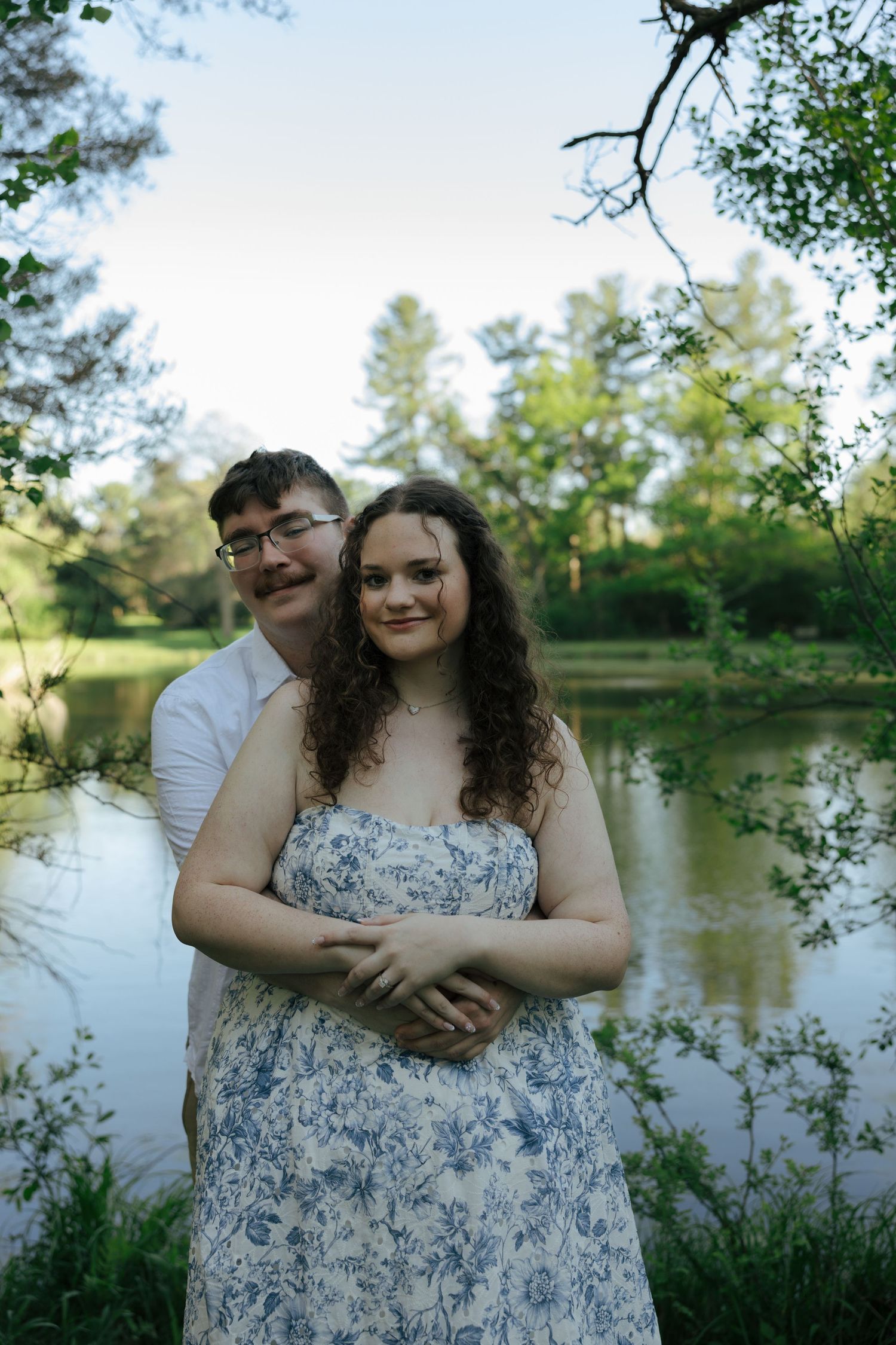 Couple embracing by a serene pond surrounded by lush green trees, woman in floral dress smiling warmly.