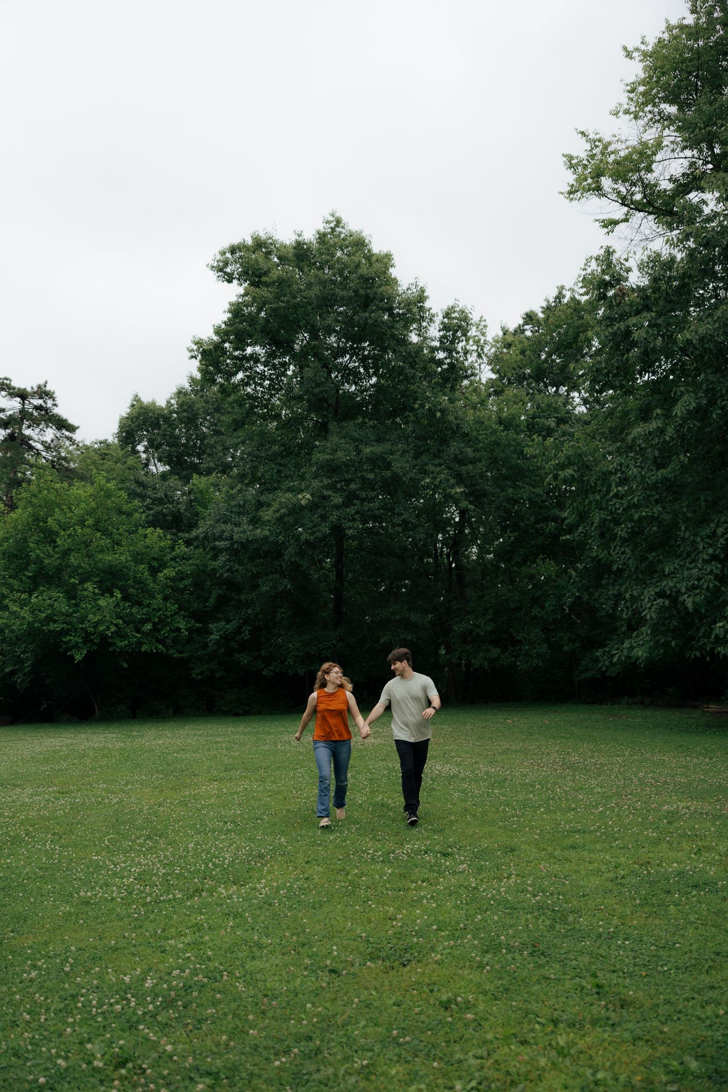 Couple strolling hand in hand through a lush green park meadow surrounded by tall trees on a cloudy day.
