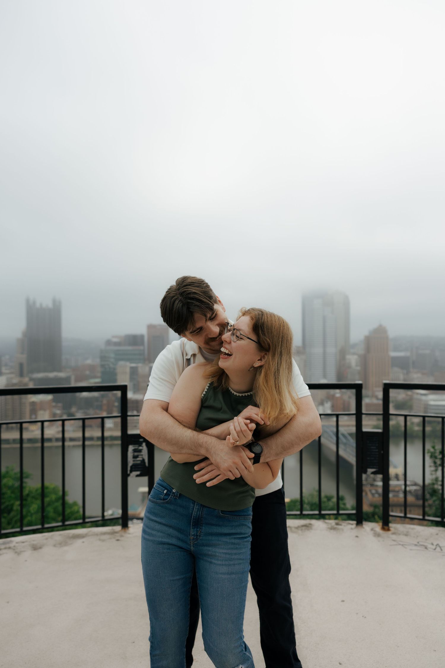 Couple embracing and laughing on a rooftop overlook with misty Pittsburgh city skyline in the background.