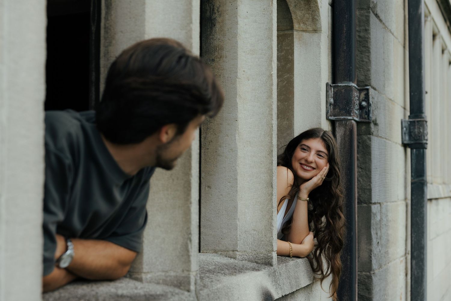 A smiling woman with long wavy hair rests her chin on her hand while a man leans nearby between stone columns.