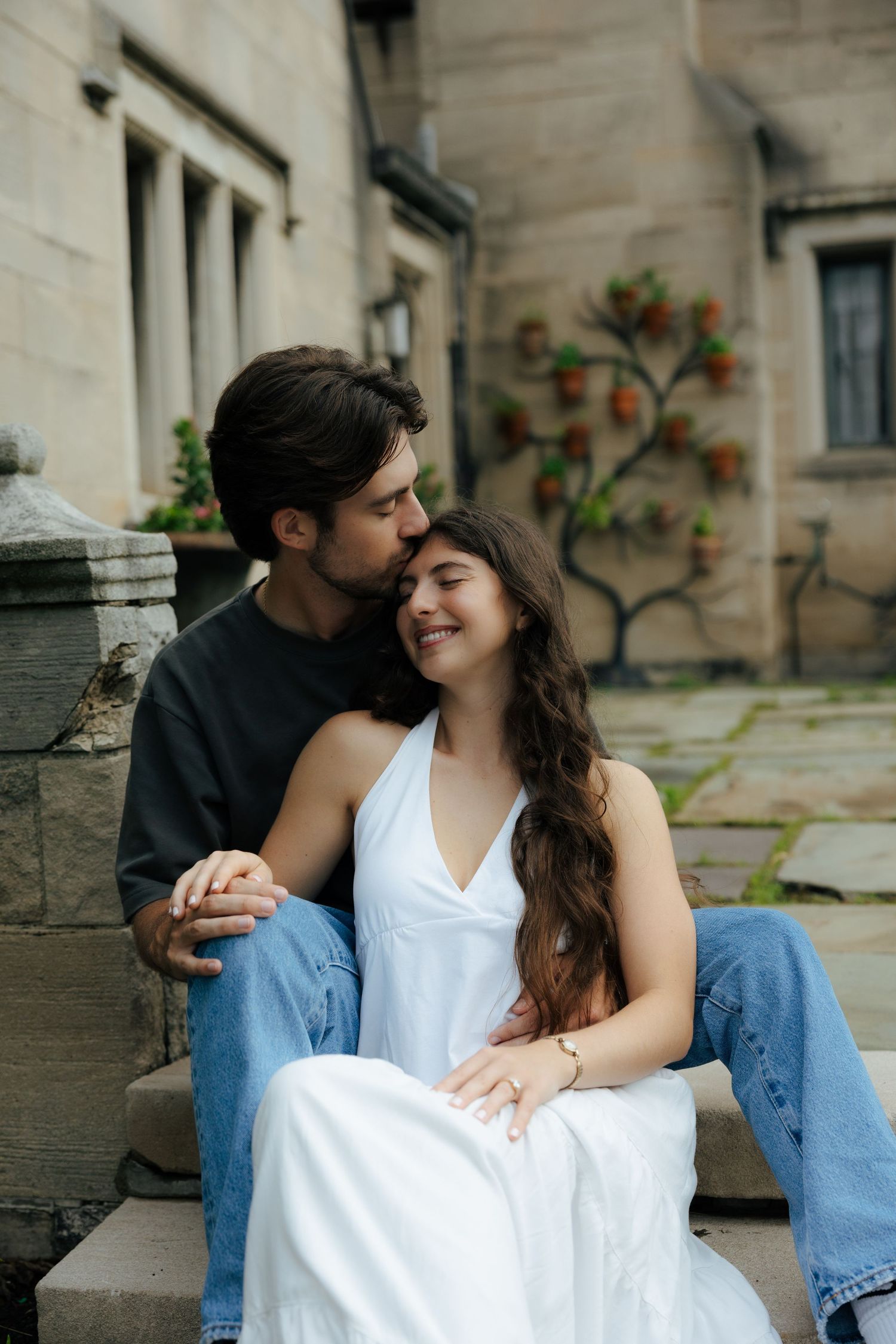 Couple sitting on stone steps embracing, man kissing womans cheek, she wears white dress.