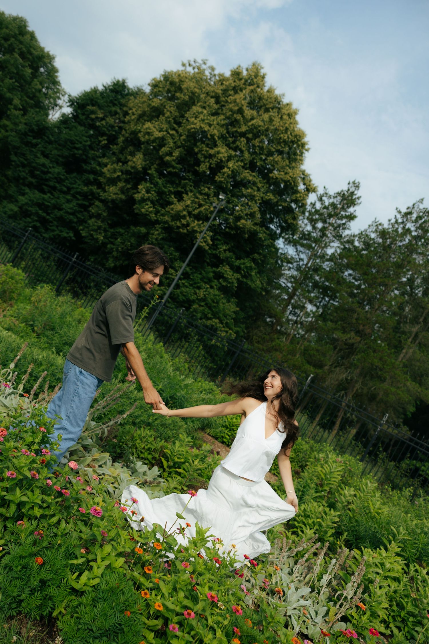 Couple holding hands in lush garden, woman in white dress laughing as man leads her through flowers.