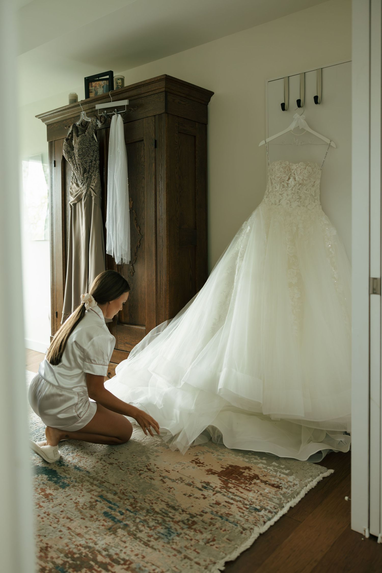 Bride kneeling on a rug adjusting her white lace ballgown wedding dress hanging on a wall hook in a rustic bedroom.