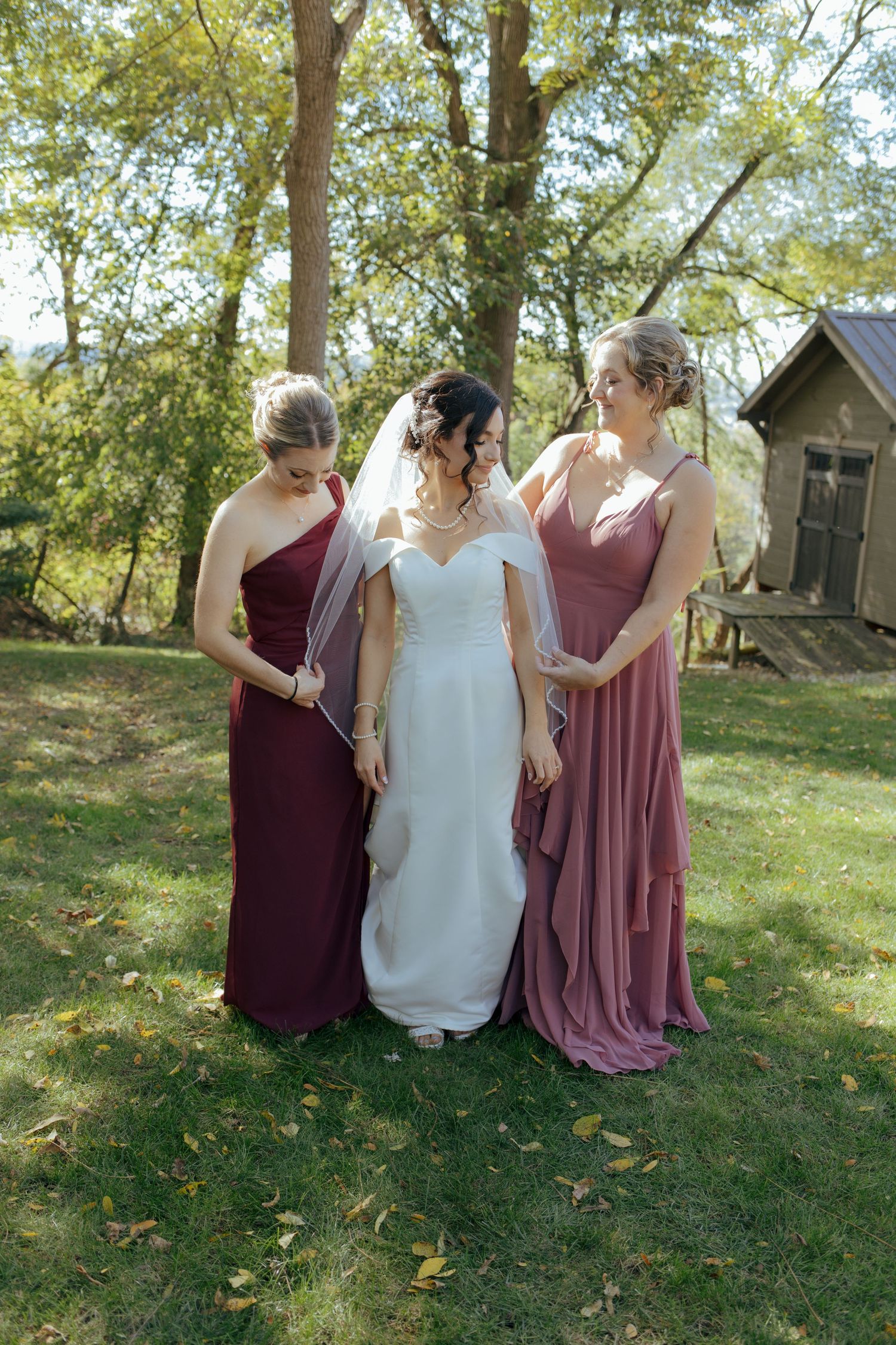 Bride in white wedding gown flanked by two bridesmaids in burgundy and mauve dresses outdoors.